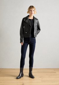 Jeune femme aux cheveux blonds courts portant une veste en cuir noir, un haut noir, un jean skinny sombre et des bottines noires, se tenant sur un sol en bois.