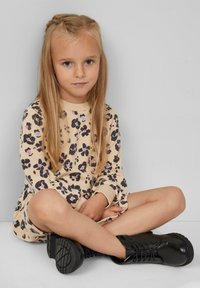 Beiges Kleid mit schwarzem und pastellfarbenem Blumenmuster, langen Ärmeln, gerippter Halslinie, kombiniert mit schwarzen Schnürboots. Sitzende Pose vor einem grauen Hintergrund.