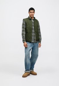 Green quilted vest over a plaid shirt, paired with light blue denim jeans and tan shoes, displayed against a plain white background.