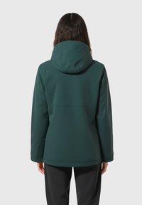 Frau mit langen Haaren, die eine dunkelgrüne Kapuzenjacke und schwarze Hosen trägt, von hinten vor einem schlichten, hellen Hintergrund gezeigt.