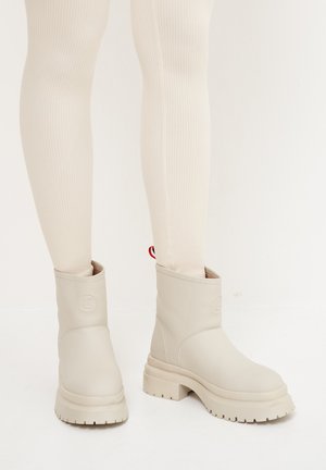 Bottines beiges en matériau synthétique lisse, avec une semelle épaisse et un discret détail de logo sur le côté. Leggings beiges fins à côtes.