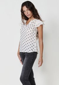 Blusa di tessuto bianco con pois neri, scollo a V, maniche corte a volant. Indossata con jeans fitted grigio scuro, che mettono in mostra un fit rilassato.
