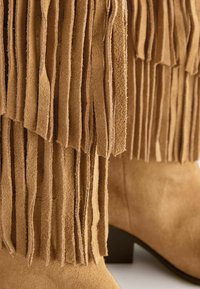 Tan suede laarzen met gelaagde franjes en lage zwarte hakken, weergegeven in close-up.