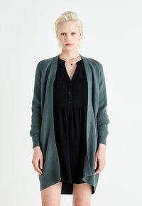 Long, open-front cardigan in dark green, made of soft, textured fabric, worn over a black dress with a buttoned neckline and flowing silhouette.