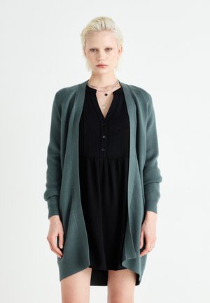 Long, open-front cardigan in dark green, made of soft, textured fabric, worn over a black dress with a buttoned neckline and flowing silhouette.