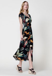 Vestido maxi con estampado floral, escote en V profundo, mangas cortas y abertura lateral; fondo negro con patrones de hojas coloridas. Lleva sandalias de cuña.