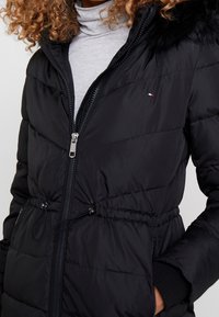 Chaqueta acolchada negra con cremallera, con capucha de piel sintética, cintura elástica y bolsillos laterales. Textura suave y patrón acolchado visible.