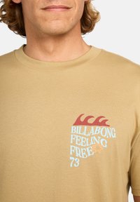 Beige langermet t-skjorte laget av mykt stoff, med en rød bølge og blå tekst "BILLABONG FEELING FREE 73" på venstre bryst.
