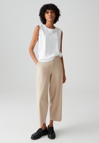 Weißes ärmelloses Oberteil, beigefarbene weite Hose, schwarze Schuhe. Die Hose verfügt über seitliche Taschen und eine subtile Textur. Einfaches, sauberes Design.