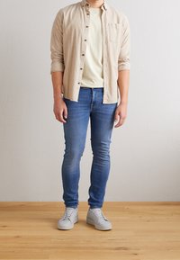 Camisa de botones beige claro sobre una camiseta amarilla clara; vaqueros ajustados azules; zapatillas grises. Tela suave, diseño entallado, look casual.
