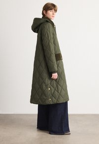 Groene, gewatteerde lange jas met capuchon, voorzien van gouden knopen, een zijzak met een donkere accenten en een relaxte silhouet over donkere wijde spijkerbroeken.