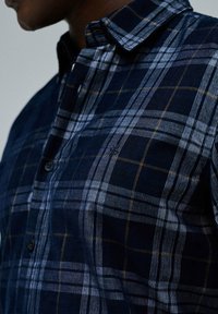 Camisa de cuadros azul marino con líneas blancas y amarillas, hecha de una tela texturizada, con cuello abotonado y botones negros.