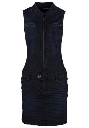 Femme portant une robe en jean noire sans manches avec fermeture éclair, des collants noirs transparents et des bottines noires, debout avec une jambe croisée.