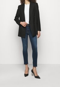 Svart dubbelknäppt blazer över en vit t-shirt, matchad med blå skinny jeans och svarta spetsiga klackar. Slät tygkvalitet och strukturerad passform.