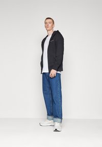 Felpa nera con zip sopra una maglietta bianca, abbinata a jeans in denim azzurro chiaro arrotolati alle caviglie e sneakers bianche con dettagli grigi.