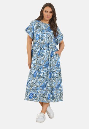 Frau in einem blau-weiß geblümten Midi-Kleid mit Rüschenärmeln und weißen Sneakers, die mit einer Hand in der Tasche steht.