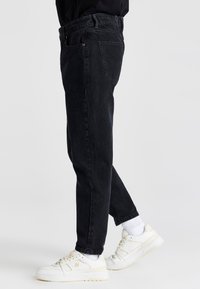 Vaqueros de mezclilla negra con un corte clásico, que cuentan con cinco bolsillos, bucles para cinturón y detalles de costura sutiles. Combinados con zapatillas blancas.