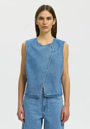 Jeune femme aux cheveux bruns raides portant un gilet asymétrique bleu en denim sans manches et un pantalon en denim taille haute assorti, sur fond blanc.