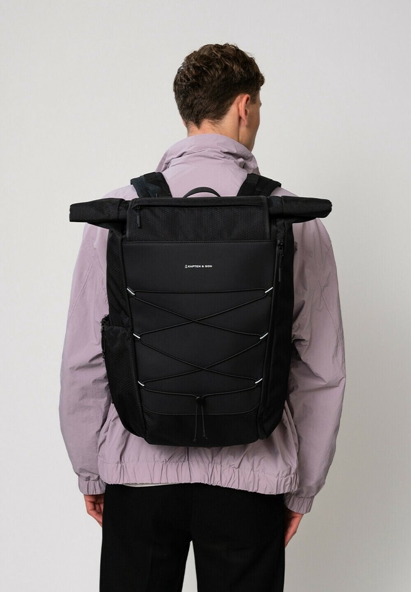 Personne portant un sac à dos noir à rabat enroulable avec cordon ajustable et poches latérales, vêtue d'une veste violet clair sur un fond uni.