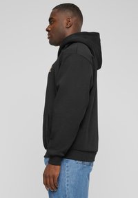 Black cotton hoodie featuring a kangaroo pocket, ribbed cuffs and hem, and a gold logo detail on the chest. Model shown from the side.