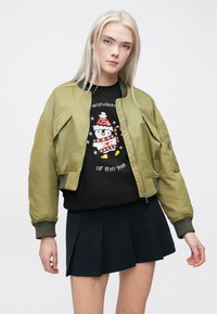 Oliven grønn bomberjakke med glidelås foran, ribbede mansjetter og to sidelommer, båret over en svart genser med brodert pingvindesign.