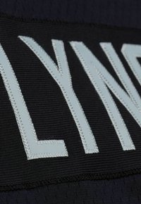 Schwarzer Stoff mit erhabenen, hellblau gestickten Buchstaben "LYN". Die Struktur wirkt gerippt und betont die Buchstabenkonturen.