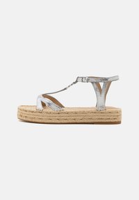Silver metallisk T-strap sandal med spänne, texturerad ovandel och tjock flätad jutesula mot en vit bakgrund.