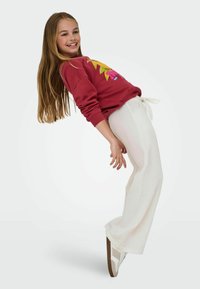 Crvena dukserica s žutim grafičkim printom, bijele teksturirane široke hlače s vezicom, i bijele tenisice s smeškastim naglascima. Uspravni stav, razigran stav.