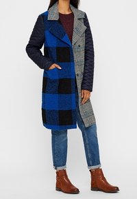 Donna che indossa un cappotto lungo a fantasia mista con un motivo a quadri blu e nero, pied-de-poule e maniche trapuntate color navy, abbinato a jeans blu e stivaletti marroni.