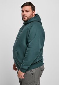 Sudadera con capucha de un verde oscuro que cuenta con un bolsillo frontal, cordones ajustables, puños de canalé y un corte relajado. La tela parece suave y texturizada.
