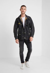 Black waxed jacket with gold buttons, plaid lining, two front pockets, and a snap closure. Paired with dark jeans and white sneakers.