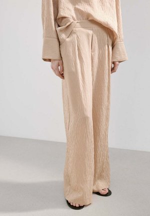 Beżowe szerokie spodnie sztruksowe noszone z dopasowanym topem, stojące boso na jasno szarej podłodze na tle jednolitej ściany.