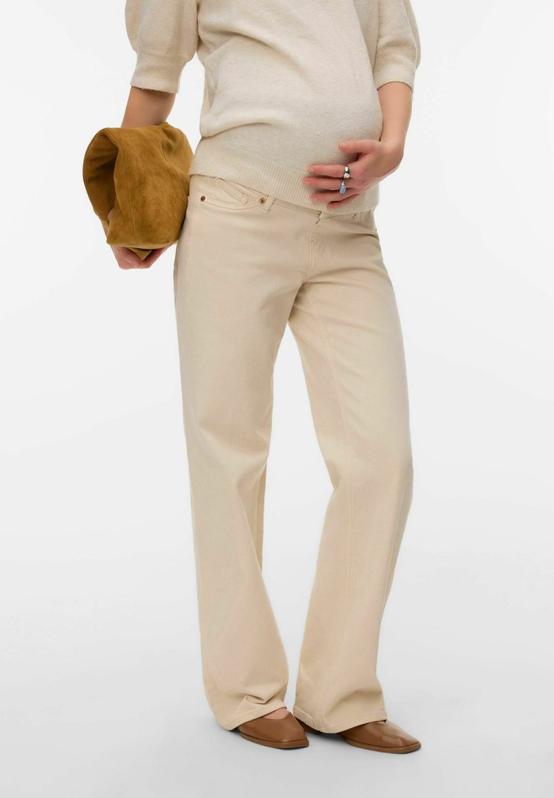 Personne enceinte en pull beige et pantalon, tenant un sac marron dans la main gauche et touchant son ventre de la main droite, portant des chaussures marron.