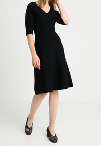 Robe en tricot noir à côtes avec décolleté en V et manches courtes, évasée à la taille. Portée avec des chaussures à talons bloc perforées noires.
