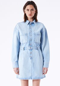Lys blå denim skjortekjole med lange ærmer, knaplukning foran, to brystlommer og en tætsiddende talje. Glat tekstur med en lige kant.