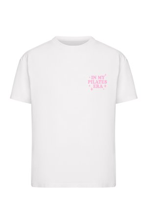 Biały t-shirt z krótkim rękawem z różowym napisem „IN MY PILATES ERA” otoczonym małymi błyskotkami na lewej stronie klatki piersiowej.