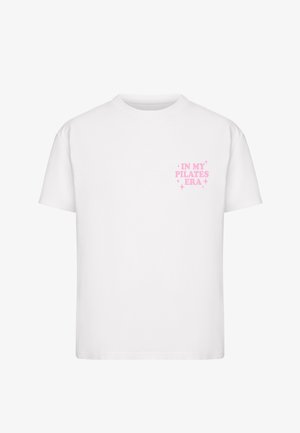 T-shirt blanc à manches courtes avec un texte rose indiquant « IN MY PILATES ERA » entouré de petites étincelles sur la zone gauche de la poitrine.