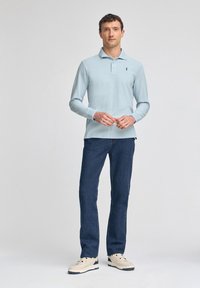 Ljusblå långärmad polo med krage och diskret logotyp, kombinerad med mörkblå jeans och beige sneakers, stående framför en neutral bakgrund.