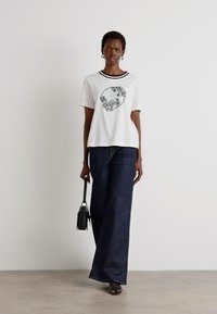 T-shirt grafica bianca con design floreale, accenti neri, abbinata a jeans larghi scuri. La modella tiene una piccola borsa nera. Sfondo minimalista.