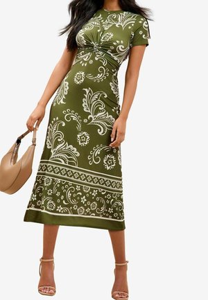 Kvinde iført olivengrøn midi-kjole med hvide blomster- og paisleymønstre, holder en beige håndtaske og bærer nøgne strop-sandaler med hæl.