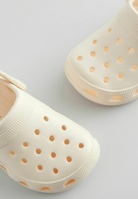 Zuecos de color beige claro, fabricados en material suave, que cuentan con múltiples agujeros de ventilación y un borde texturizado, además de una correa trasera para mayor soporte.