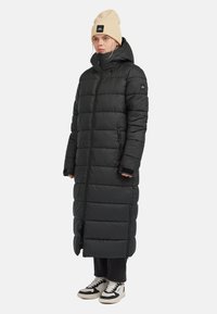  Lange zwarte pufferjas met een hoge kraag, gevoerde secties, ritszakken en geribbelde mouwen. Draagt over zwarte broek en sneakers met witte accenten.
