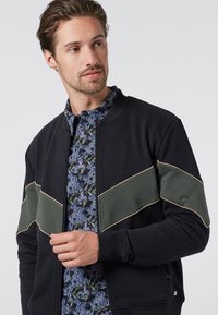 Felpa nera con zip con strisce orizzontali verdi e contorno beige. Indossata sopra una camicia a fantasia floreale, che mette in mostra un look a strati.