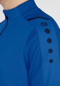 Giacca sportiva blu con colletto alto, chiusura con zip, tessuto leggero, finitura testurizzata e accenti neri, tra cui dettagli circolari sulle spalle.