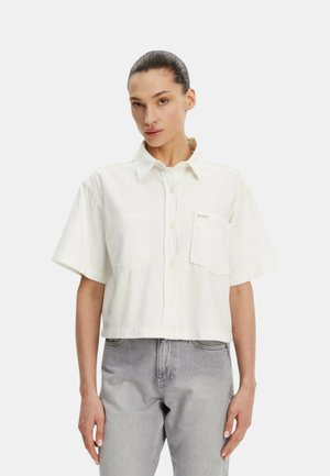 Donna con camicia bianca a maniche corte con tasca frontale, abbinata a jeans grigio chiaro a vita alta, in piedi davanti a uno sfondo semplice.