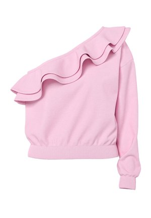 Lyshåret kvinde stående med hænderne bag ryggen iført en pink asymmetrisk rynket top, lyse denimshorts og sorte flipflops.