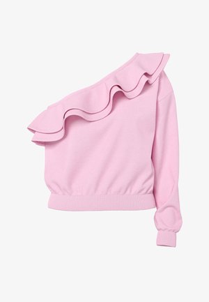 Lyshåret kvinde stående med hænderne bag ryggen iført en pink asymmetrisk rynket top, lyse denimshorts og sorte flipflops.