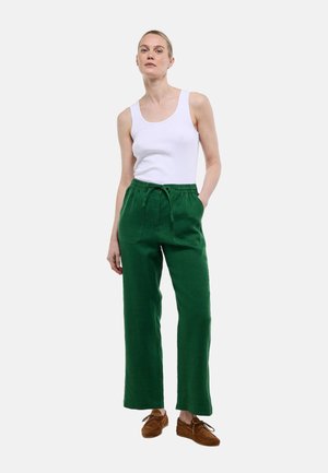 Femme portant un haut blanc sans manches, un pantalon vert avec cordon de serrage, et des chaussures marron à lacets, debout avec une main dans la poche.