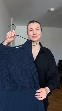 Γυναίκα με μαλλιά μαζεμένα πίσω, που φοράει μαύρο πουκάμισο, κρατάει σε μια κρεμάστρα ένα σκούρο μπλε δαντελένιο φόρεμα σε ένα απλό δωμάτιο.