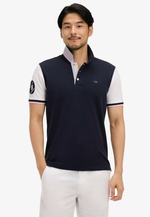 Homme portant un polo bleu marine et blanc à manches courtes avec col, et un pantalon blanc, debout avec une main dans la poche, sur fond blanc.